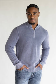 A man wearing a ribbed long-sleeve half-zip sweater in a dusty periwinkle blue color, paired with blue jeans, standing against a plain light gray background.