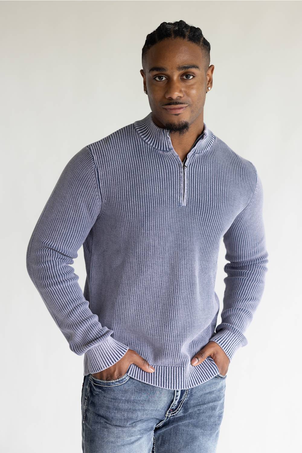 A man wearing a ribbed long-sleeve half-zip sweater in a dusty periwinkle blue color, paired with blue jeans, standing against a plain light gray background.
