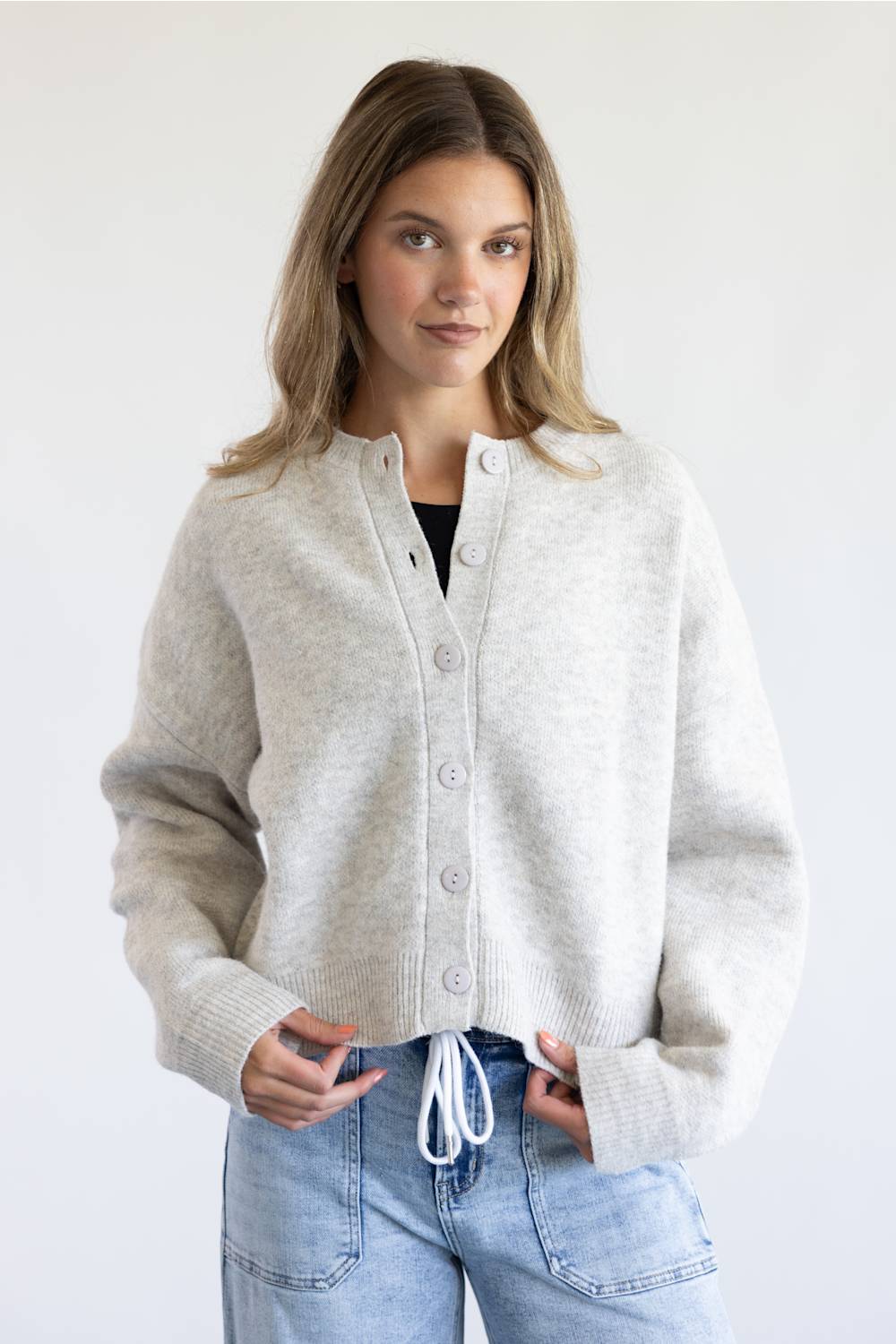 A woman modeling a light gray knit cardigan with a button-up front and drawstring detail, paired with light blue jeans against a plain white background.