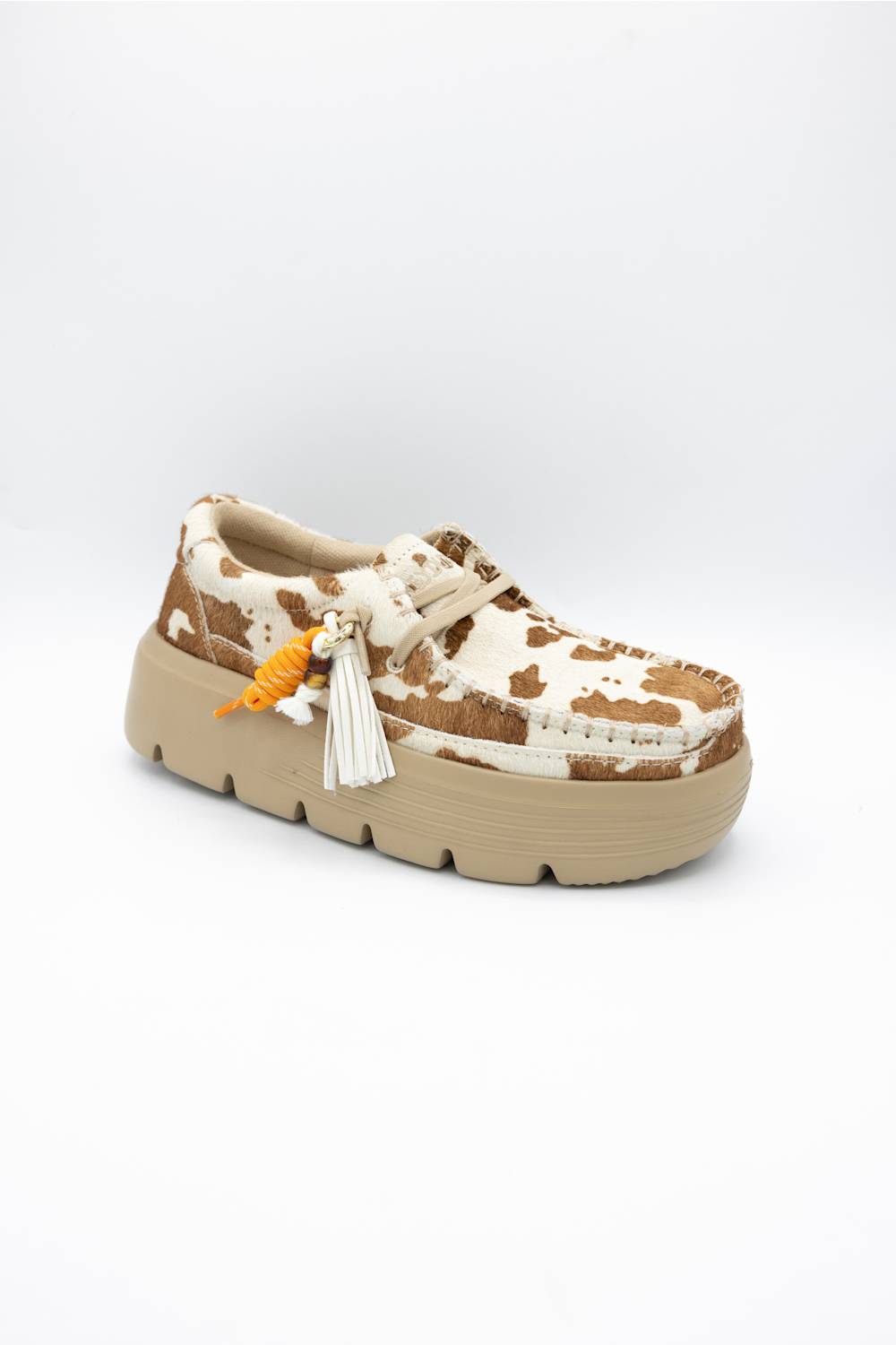 A single platform shoe with a brown and cream cow print upper, featuring a chunky beige sole, white laces, and a decorative tassel ornament.