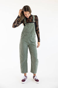 Front view of model posing with hand touching hair, wearing Aaron & Amber Slouchy Denim Overalls for Women in Olive over a black sheer floral lace long-sleeve top paired with burgundy low-top sneakers.