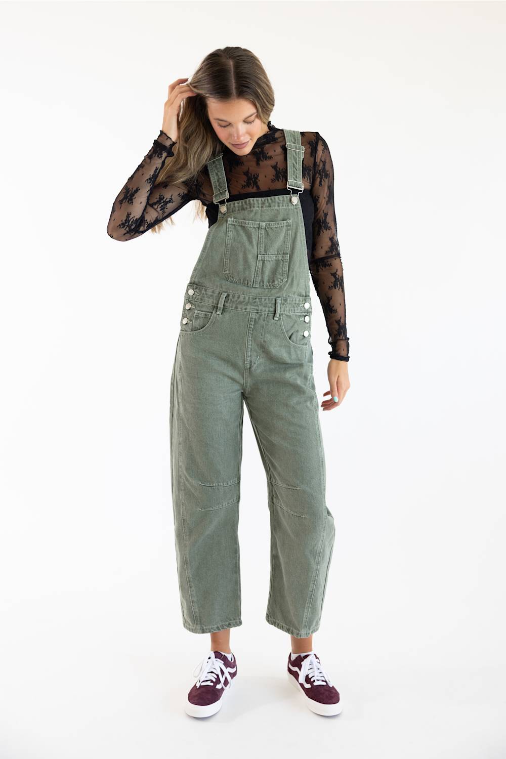 Front view of model posing with hand touching hair, wearing Aaron & Amber Slouchy Denim Overalls for Women in Olive over a black sheer floral lace long-sleeve top paired with burgundy low-top sneakers.