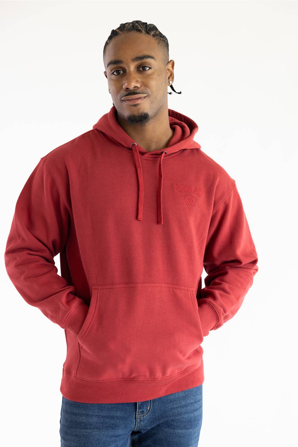 A man wearing a red pullover hoodie with a tonal embroidered chest logo, paired with blue jeans, photographed against a plain white studio background.