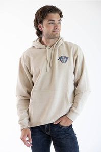 A man wearing a beige pullover hoodie with a blue embroidered 'Ariat' logo on the left chest, paired with dark blue jeans, photographed against a clean white studio background.