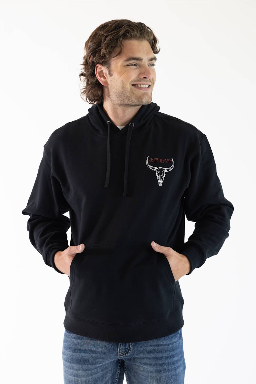 A man wearing a black pullover hoodie with an Ariat bull logo on the left chest, paired with blue jeans, photographed against a plain white background.