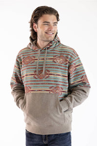 A man wearing a patterned pullover hoodie with a solid tan lower body and front pouch pocket, paired with dark blue jeans, against a plain white background.
