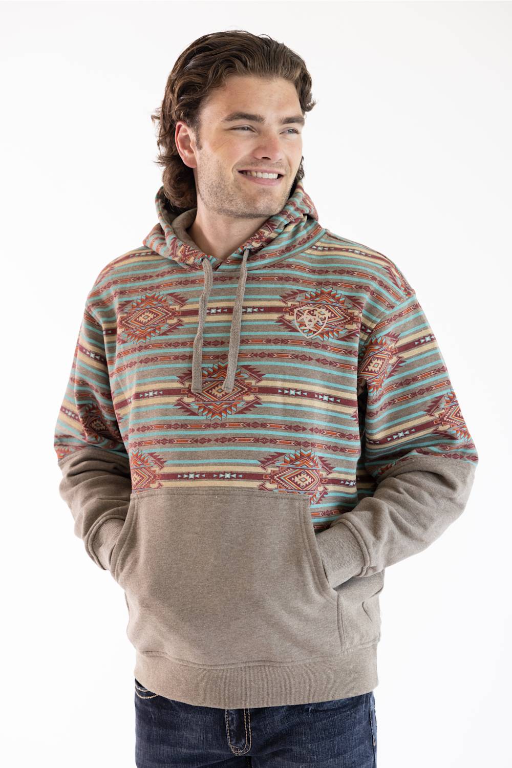 A man wearing a patterned pullover hoodie with a solid tan lower body and front pouch pocket, paired with dark blue jeans, against a plain white background.