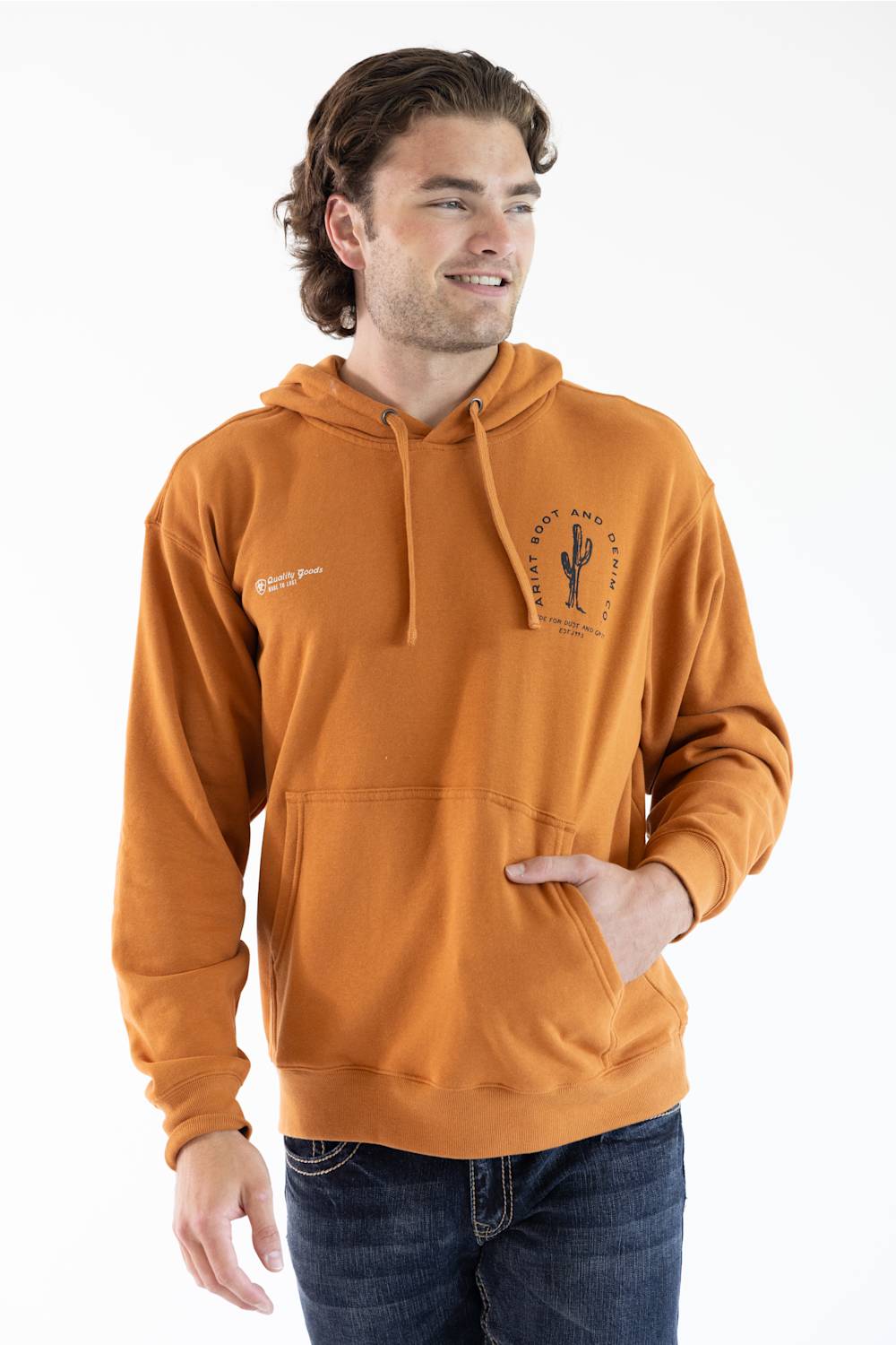 A man wearing a burnt orange pullover hoodie with a drawstring hood and kangaroo pocket, featuring a black cactus graphic on the left chest and a small white graphic on the right chest, paired with dark blue jeans against a white studio background.