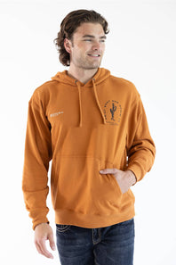 A man wearing a burnt orange pullover hoodie with a drawstring hood and kangaroo pocket, featuring a black cactus graphic on the left chest and a small white graphic on the right chest, paired with dark blue jeans against a white studio background.