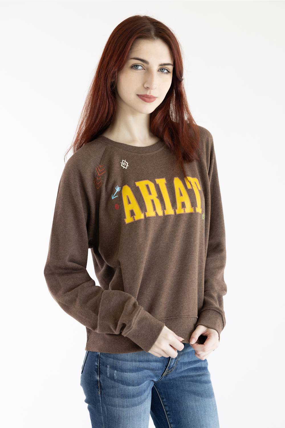 A woman with red hair wearing a brown crewneck sweatshirt with 'ARIAT' printed in yellow across the chest, paired with blue jeans, against a plain white background.
