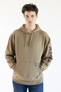 A man wearing a tan pullover hoodie with 'ARIAT' embroidered vertically in brown on the right sleeve and a small yellow logo patch near the cuff, paired with blue jeans against a plain white background.