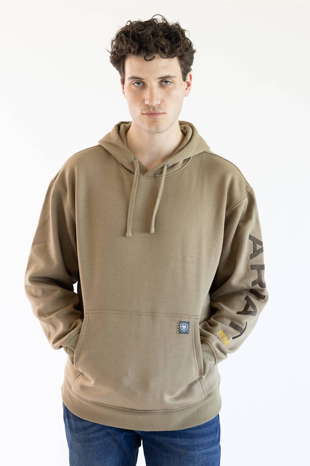 A man wearing a tan pullover hoodie with 'ARIAT' embroidered vertically in brown on the right sleeve and a small yellow logo patch near the cuff, paired with blue jeans against a plain white background.