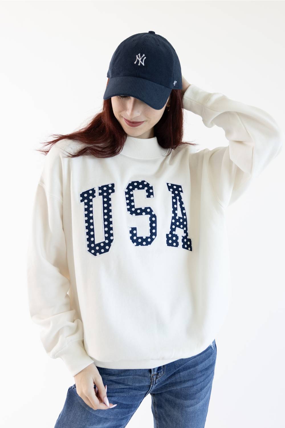 A woman wearing a cream-colored turtleneck sweatshirt with 'USA' in navy blue letters with white polka dots, paired with a navy blue baseball cap and blue jeans, against a plain white background.