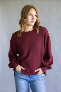 A woman wearing a maroon waffle-knit long-sleeve sweater and blue jeans, standing against a neutral gray studio background.
