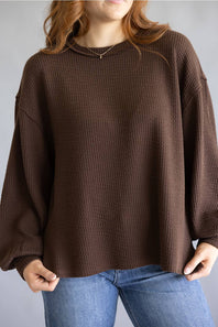 A woman wearing a brown waffle-knit long-sleeve sweater and blue jeans, shown from the neck down against a plain gray background.