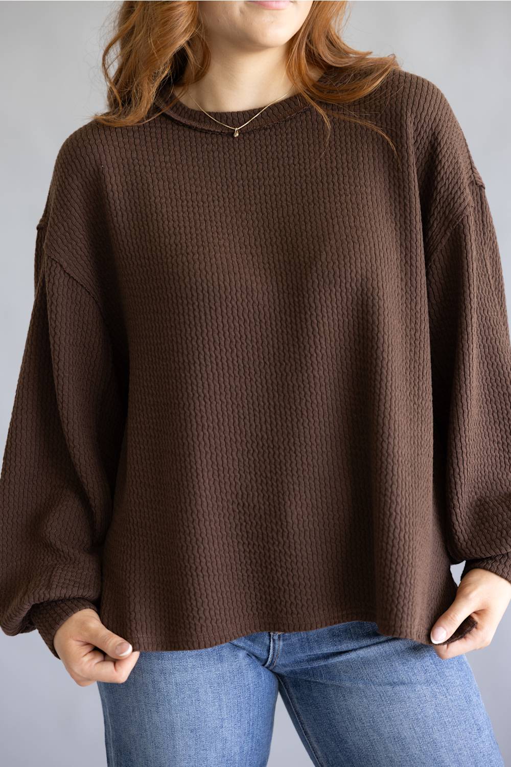 A woman wearing a brown waffle-knit long-sleeve sweater and blue jeans, shown from the neck down against a plain gray background.