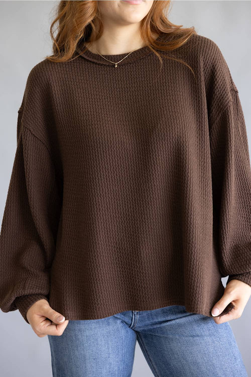A woman wearing a brown waffle-knit long-sleeve sweater and blue jeans, shown from the neck down against a plain gray background.