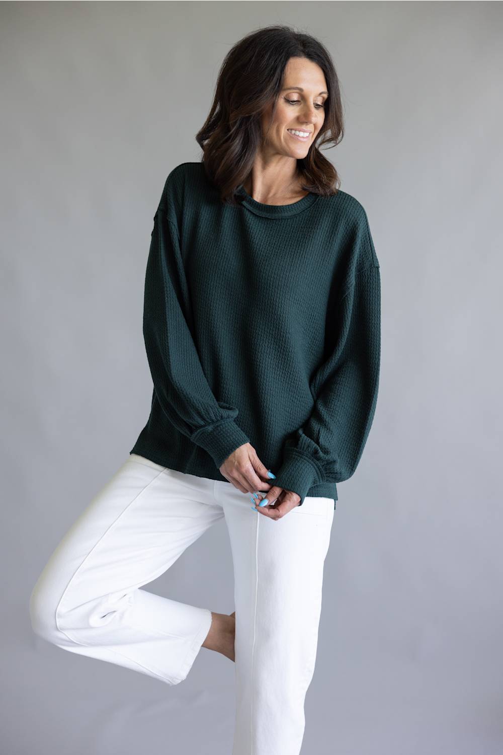A woman wearing a dark forest green ribbed knit sweater and white cropped pants, posing against a neutral gray studio background.