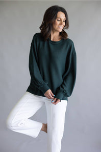 A woman wearing a dark forest green ribbed knit sweater and white cropped pants, posing against a neutral gray studio background.