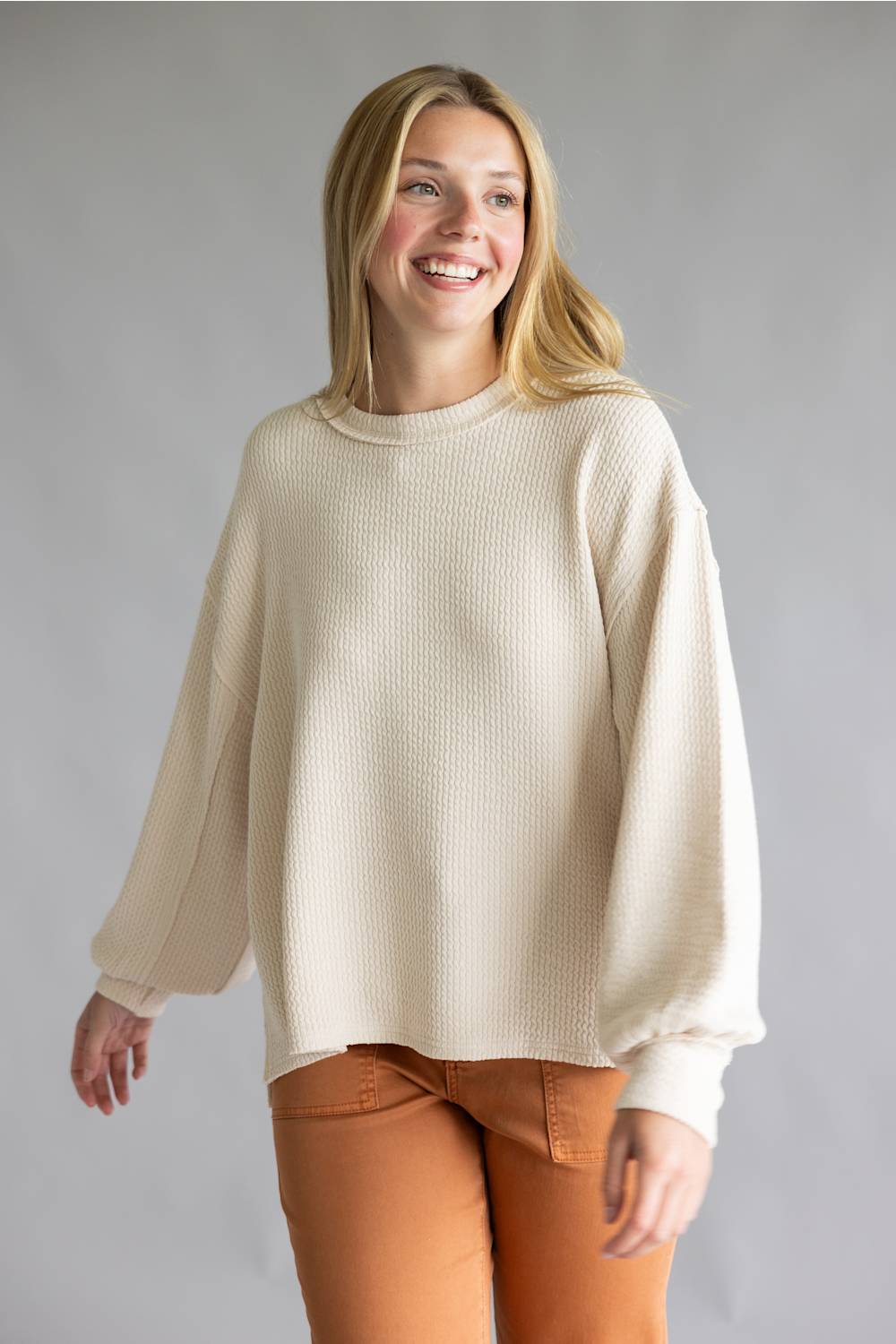 A smiling blonde woman wearing a cream-colored waffle-knit sweater and rust-orange pants, standing against a neutral gray studio background.