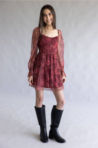 A woman modeling a short burgundy floral dress with sheer long sleeves and black knee-high boots, standing against a light gray studio backdrop.
