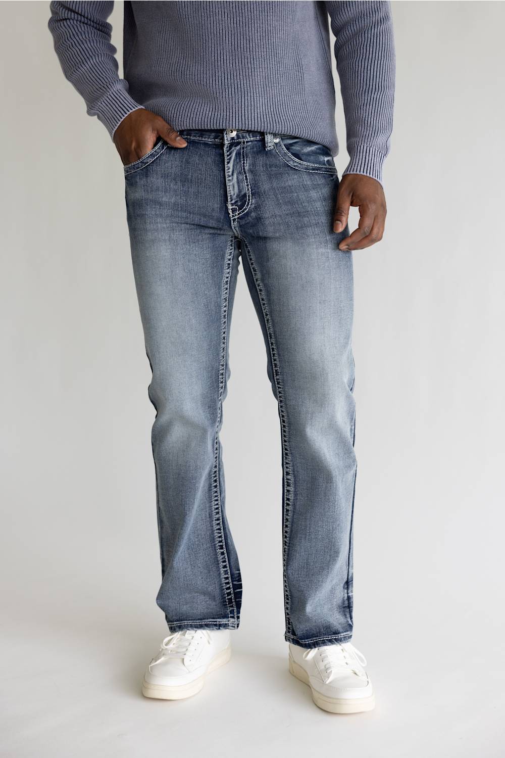 A person wearing light-wash blue jeans, a ribbed gray sweater, and white sneakers, standing against a plain light gray studio background.