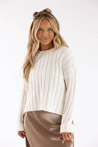 A woman modeling a cream ribbed knit long-sleeve sweater with a brown satin skirt and a brown bow headband, set against a plain white studio background.