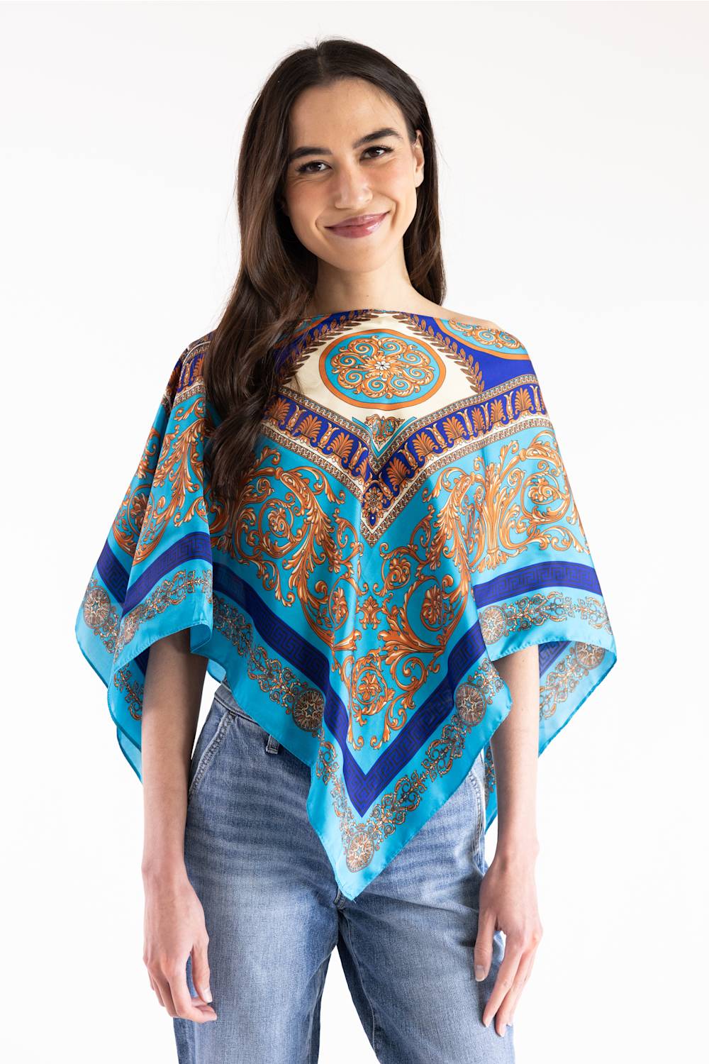 A woman modeling a vibrant paisley-patterned poncho top with blue, orange, and beige tones, paired with blue jeans, against a plain white background.