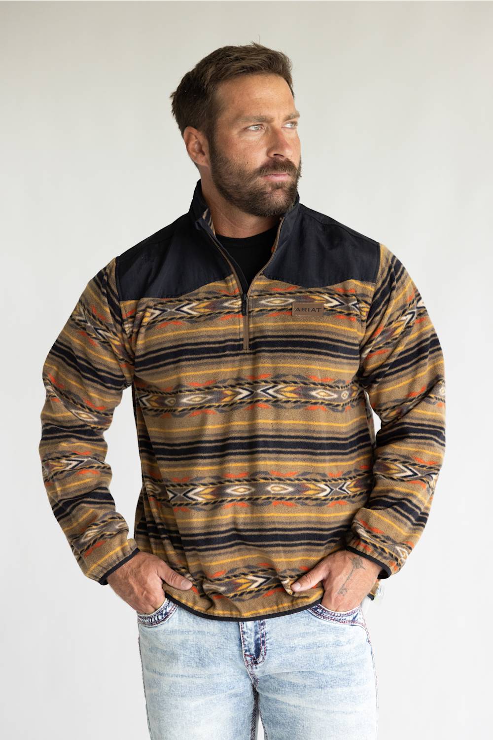 A man wearing a quarter-zip fleece pullover with a Southwestern geometric pattern in brown, tan, black, and orange, paired with light-wash jeans, photographed against a neutral light gray background.