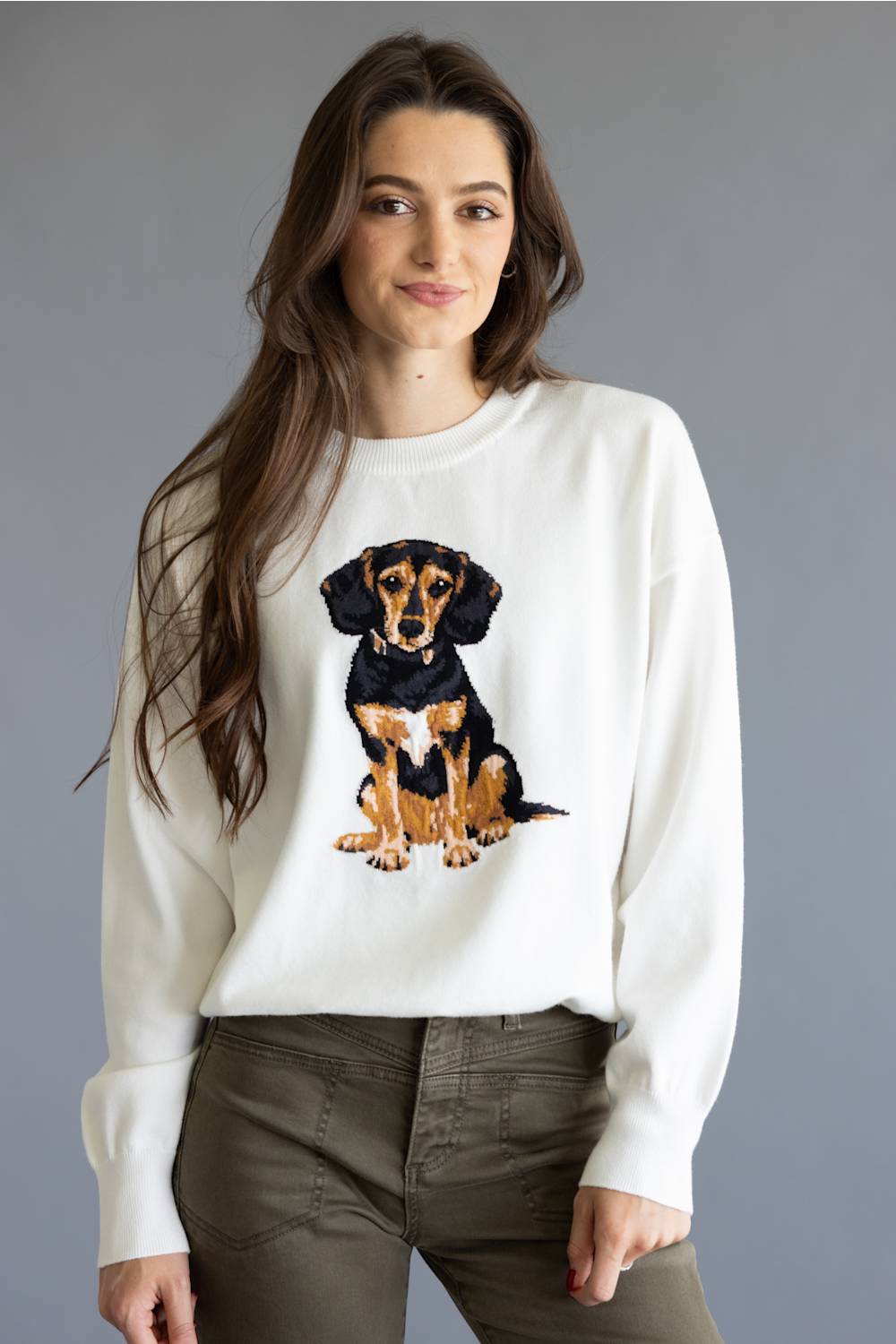 A woman wearing a white crewneck sweatshirt with a large embroidered dachshund graphic on the front, paired with olive green pants, set against a solid gray studio background.