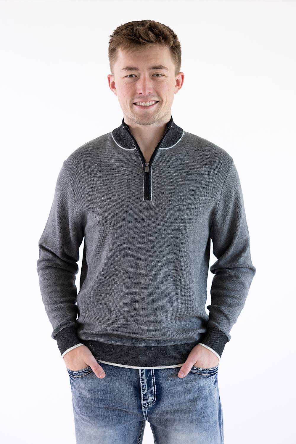 A male model facing forward wearing the F/X Fusion Birdseye Knit Quarter Zip in nickel gray, featuring a mock neck, quarter-zip placket, and dark contrasting trim along the collar, cuffs, and hem, paired with classic denim jeans.