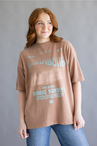 A woman with red hair wearing an oversized brown graphic t-shirt with light blue text that reads 'SUBMARINE' and 'MAGIC SUNSET UNDERWATER', paired with blue jeans, against a gray studio background.