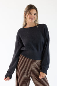 A woman modeling a dark charcoal gray knit sweater with a wide boat neckline and ribbed cuffs, paired with brown and beige striped pants, against a plain white studio background.