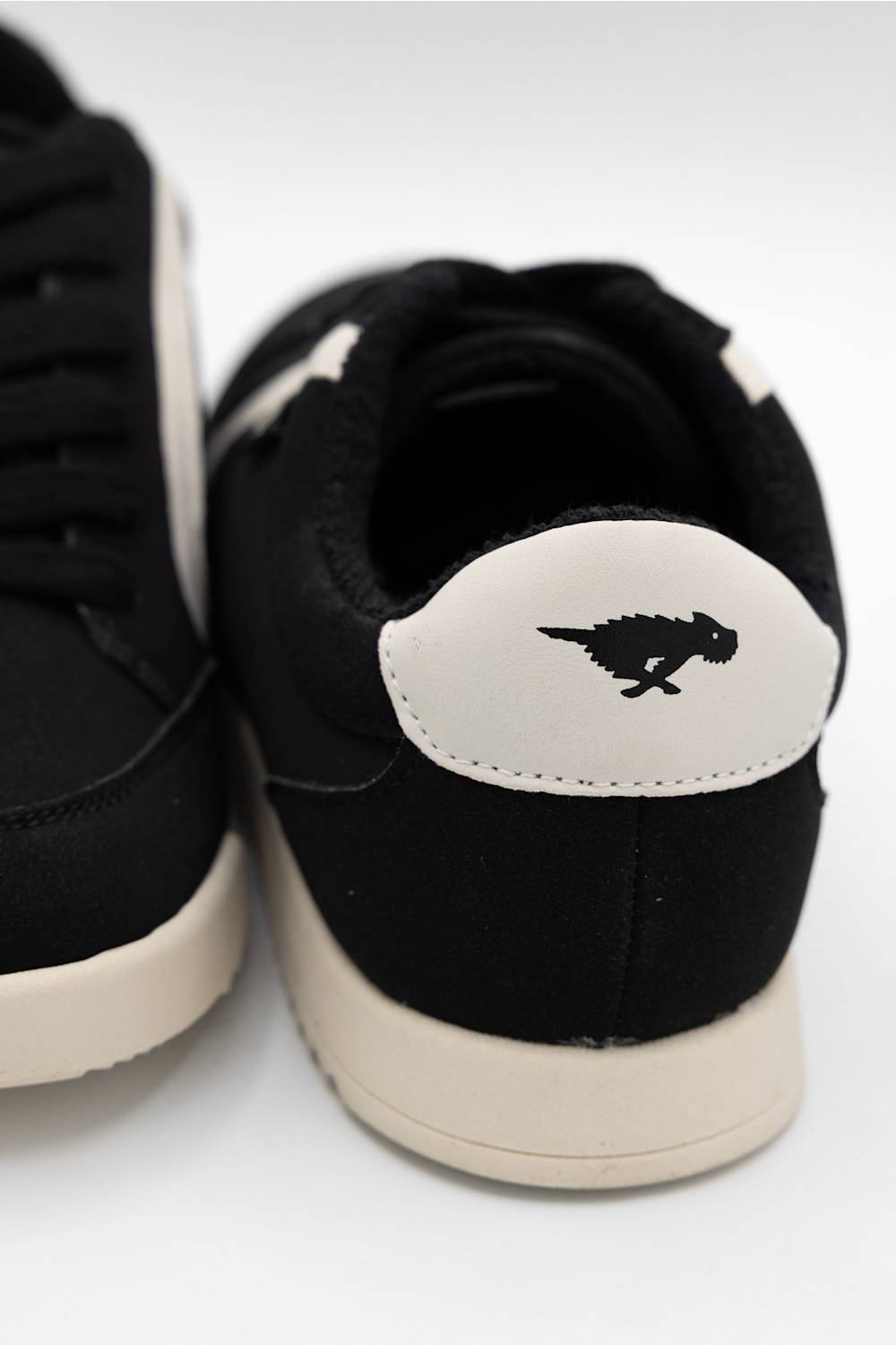 Close-up of a black low-top sneaker showing the heel and back collar, with a cream-colored heel tab featuring a black dinosaur silhouette logo and a matching cream midsole.