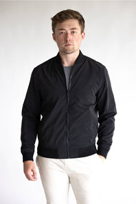 A man wearing a black zip-up bomber jacket over a dark gray t-shirt and off-white trousers, standing against a plain light gray studio background.