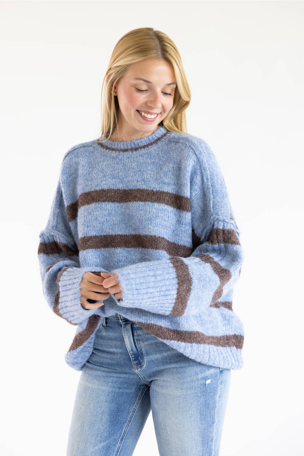 A woman wearing a light blue knit sweater with horizontal brown stripes and light-wash blue jeans, standing against a plain white studio background.