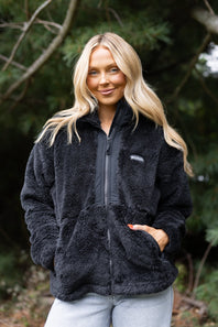 A woman with blonde hair wearing a black fleece zip-up jacket and light blue jeans, standing outdoors in front of a blurred background of trees.