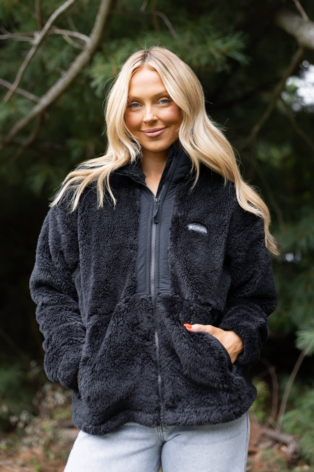 A woman with blonde hair wearing a black fleece zip-up jacket and light blue jeans, standing outdoors in front of a blurred background of trees.