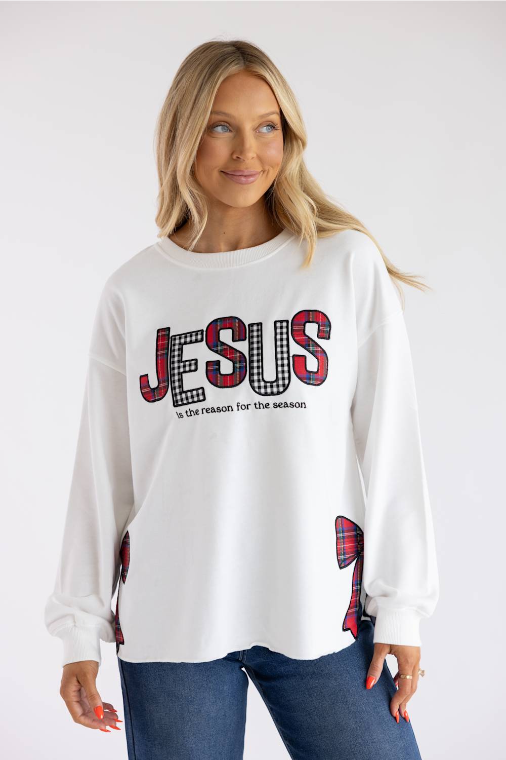 A woman wearing a white long-sleeve sweatshirt with 'JESUS is the reason for the season' printed in red and black plaid and white on the front, paired with blue jeans, against a light gray studio background.