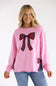 A woman wearing a pink crewneck sweatshirt with red and green plaid bow graphics on the front and sleeve, paired with blue jeans, against a plain white studio background.