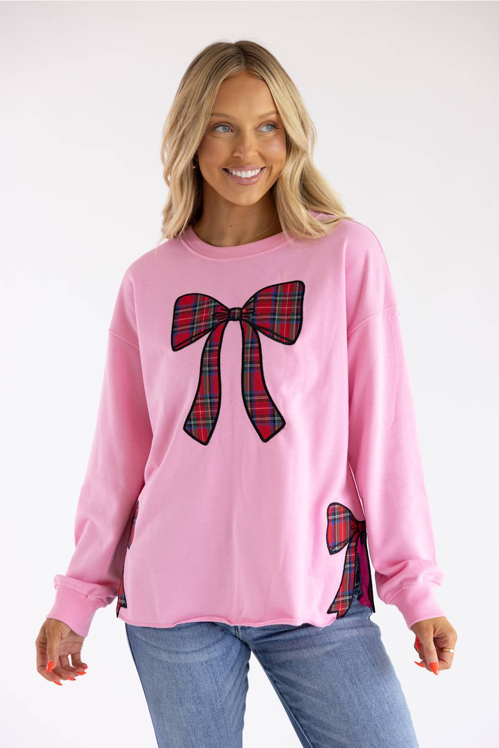 A woman wearing a pink crewneck sweatshirt with red and green plaid bow graphics on the front and sleeve, paired with blue jeans, against a plain white studio background.