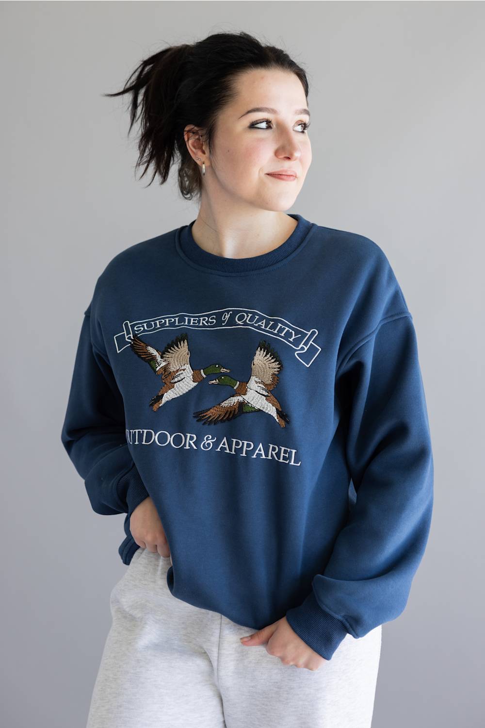 A woman wearing a navy blue crewneck sweatshirt with a graphic of two flying ducks and the text 'SUPPLIERS OF QUALITY' and 'OUTDOOR & APPAREL', paired with light gray sweatpants, against a plain gray background.