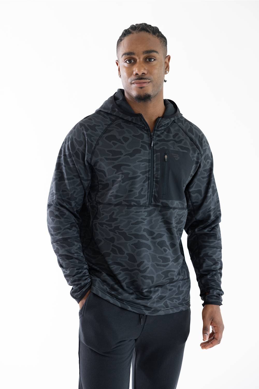A man wearing a dark gray and black camouflage patterned quarter-zip hoodie with a chest pocket, paired with black pants, photographed against a solid white studio background.