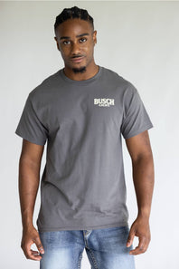 A man wearing a dark gray crewneck t-shirt with a white 'BUSCH Light' logo on the left chest, paired with blue jeans, standing against a light gray background.