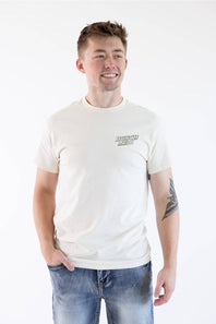 A man wearing a cream-colored short-sleeve t-shirt with a small black graphic on the left chest, paired with blue jeans, standing against a plain white background.