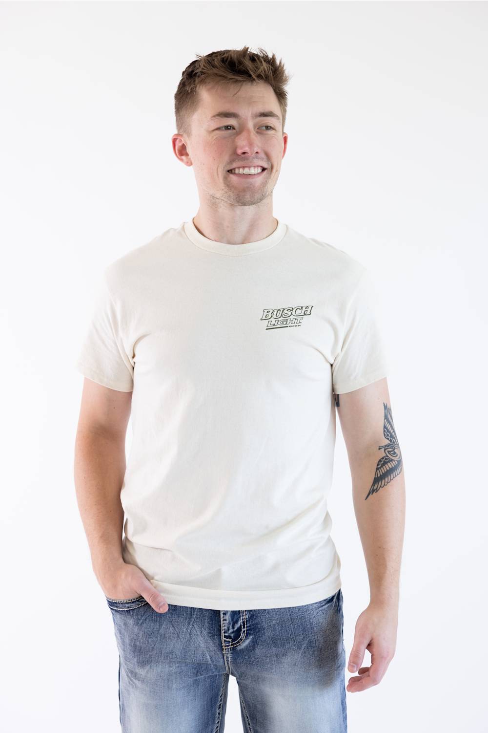 A man wearing a cream-colored short-sleeve t-shirt with a small black graphic on the left chest, paired with blue jeans, standing against a plain white background.
