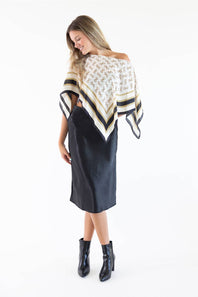 A woman modeling a cream and black patterned square scarf draped off her shoulders over a black midi skirt and black ankle boots, photographed against a plain white studio background.