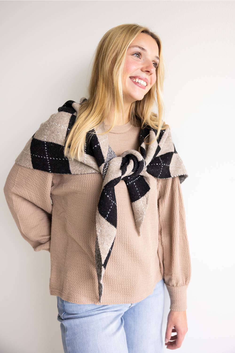 A blonde woman smiling as she wears a khaki waffle-knit crewneck sweater styled with a black and beige argyle sweater tied around her shoulders and light blue jeans against a plain white background.