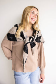 A blonde woman smiling as she wears a khaki waffle-knit crewneck sweater styled with a black and beige argyle sweater tied around her shoulders and light blue jeans against a plain white background.