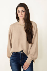 A woman with long dark brown hair wearing a beige waffle-knit long-sleeve sweater and dark blue jeans, standing against a plain off-white background.